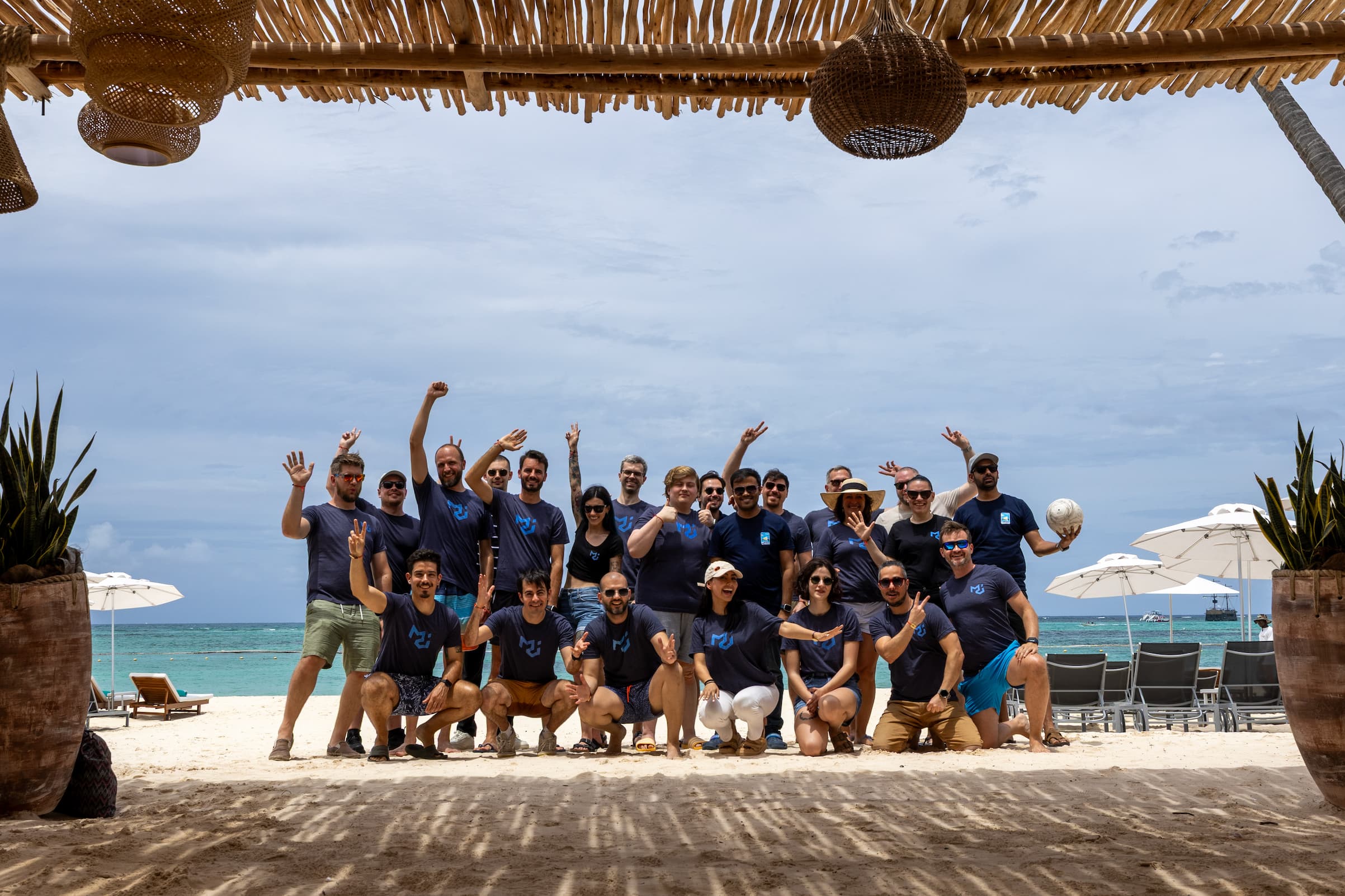 The MUI team poses together in matching dark blue t-shirts on a white sand beach with the ocean in the background.